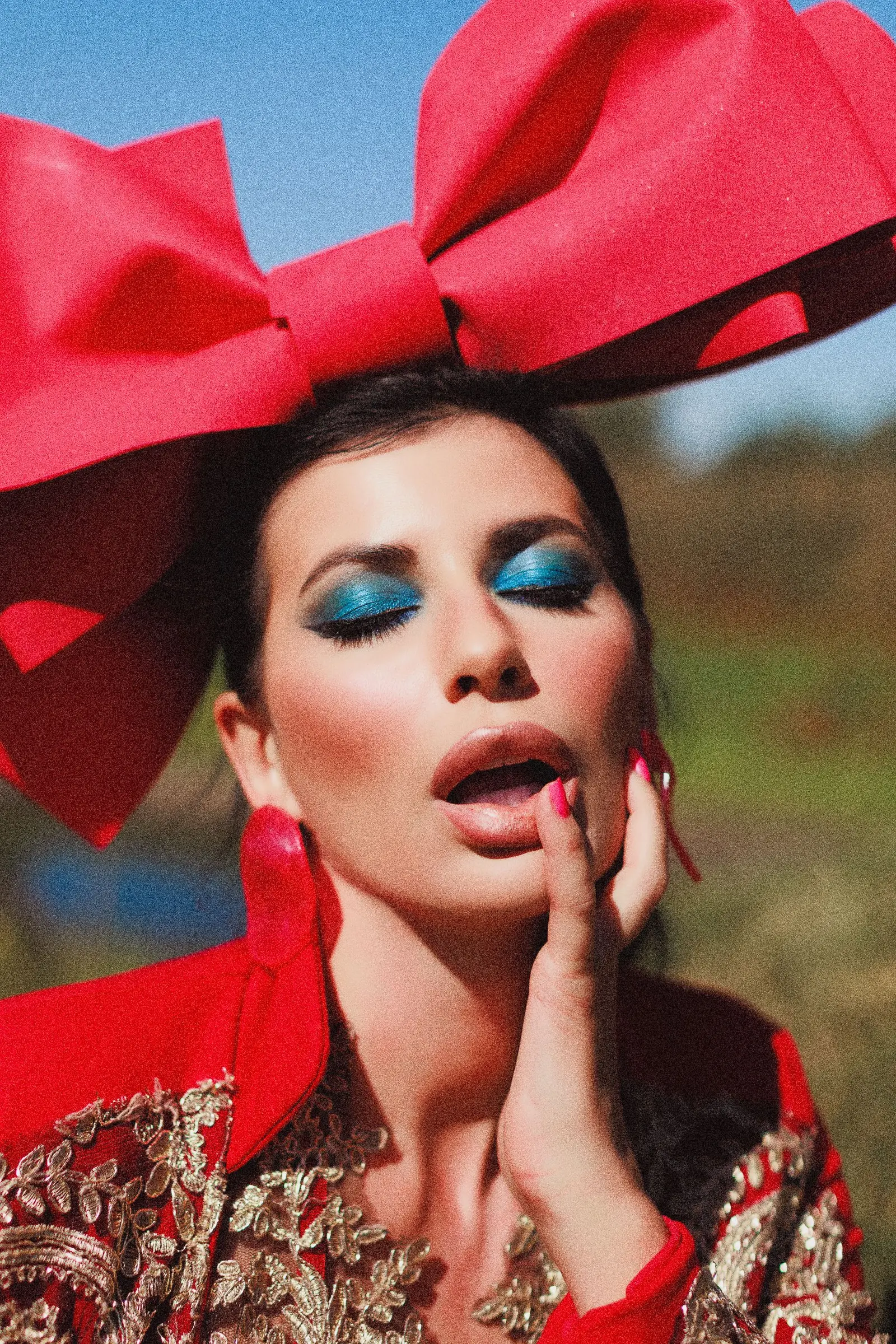 A woman in a red satin bow and red jacket with bright blue eyeshadow, her hand raised to her lips