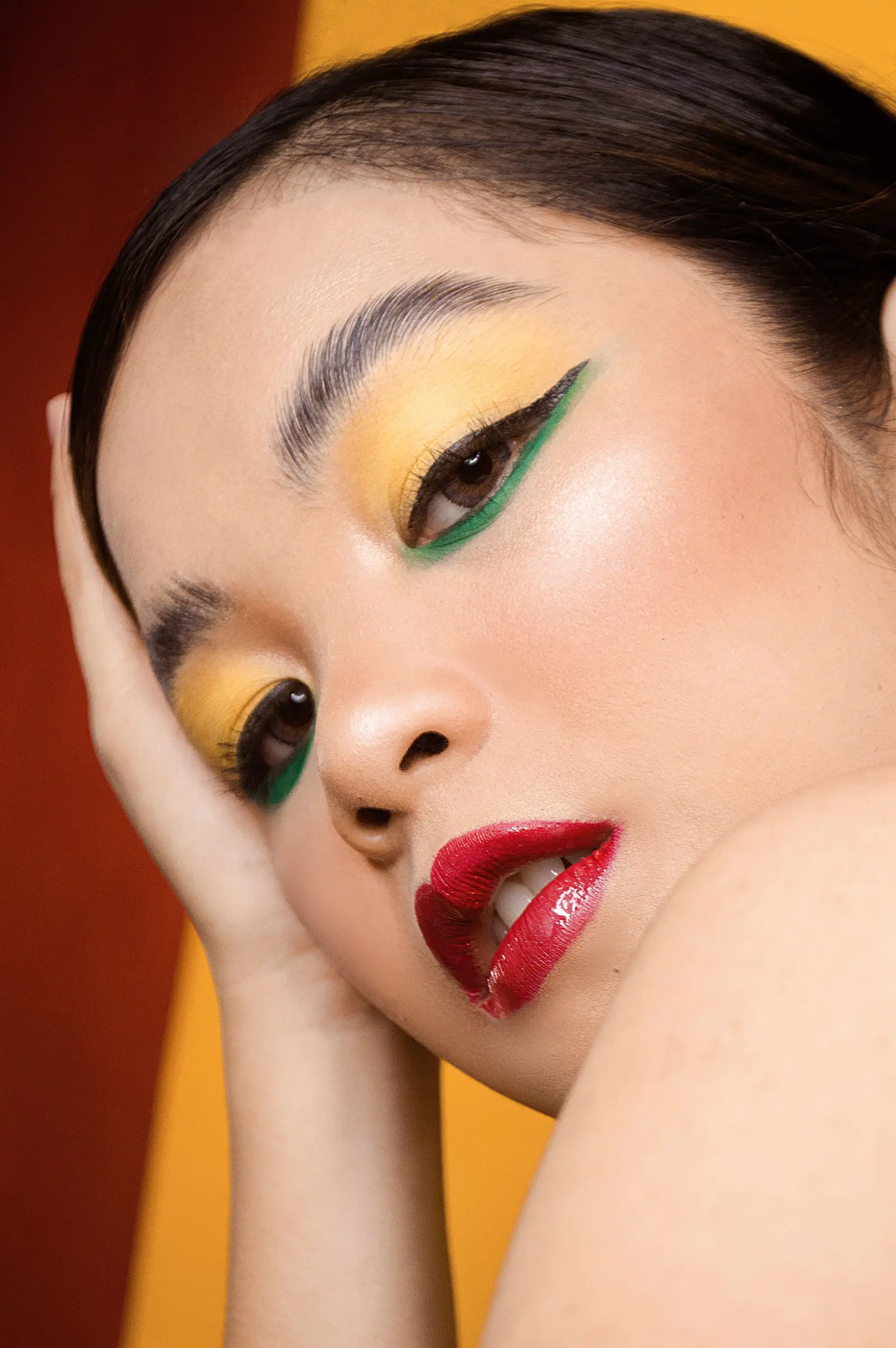 A woman with graphic yellow and green eyeliner and glossy red lips, resting her head on her hand