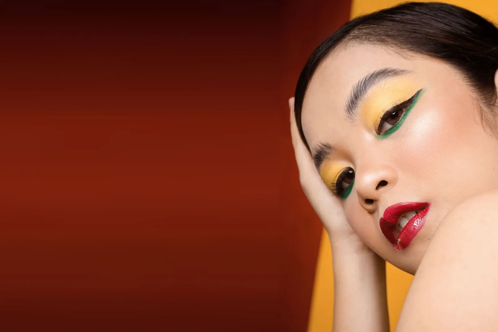 A woman with bold yellow eyeshadow, a sharp green liner flick and glossy red lips, resting her cheek on her hand against a rust-red backdrop