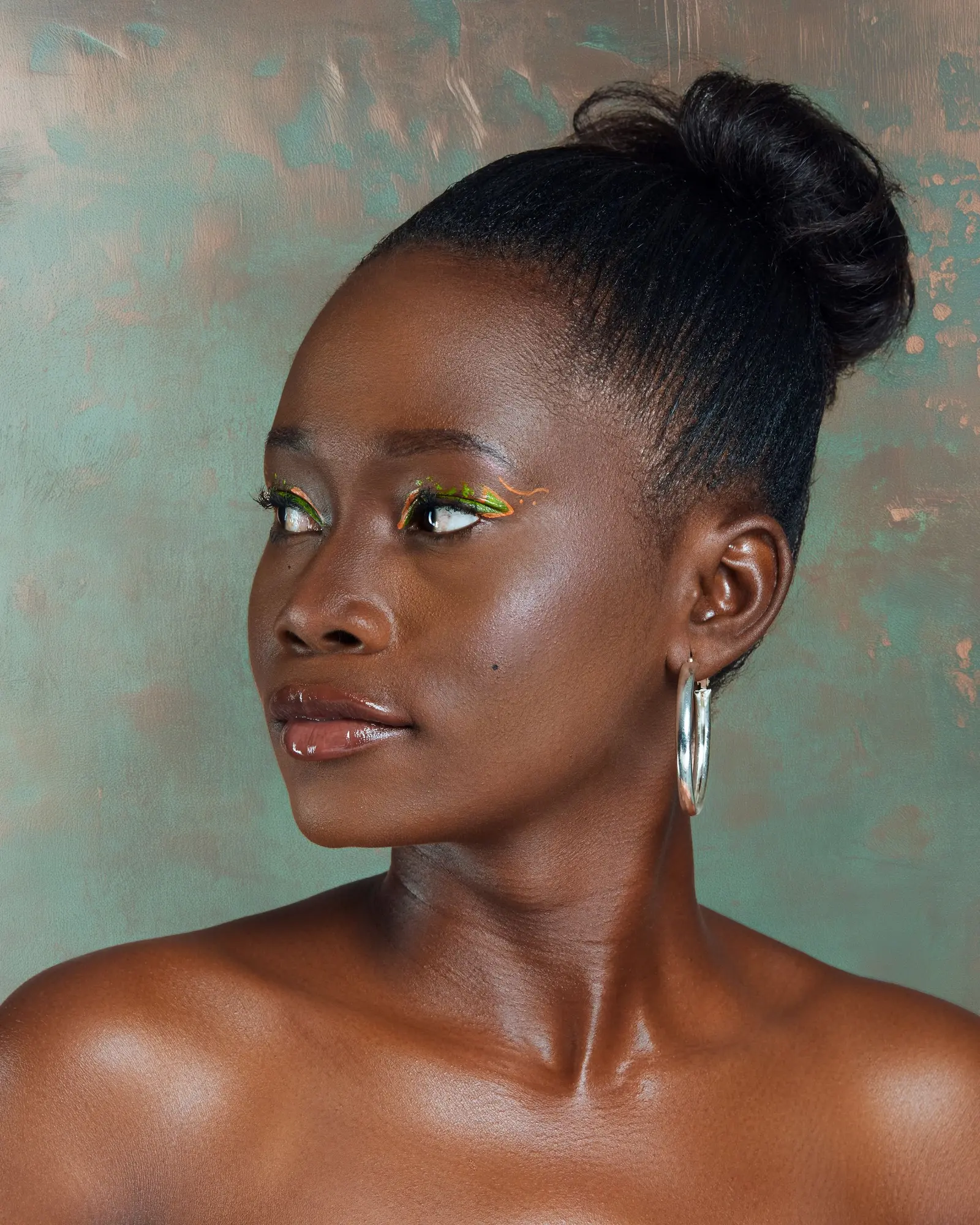 A woman with her hair in an updo, a hoop earring and a flick of green liner, against a teal wall