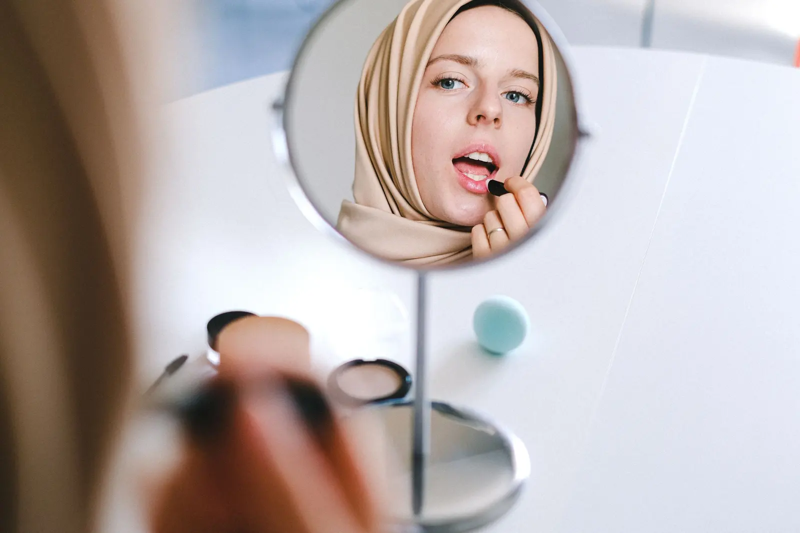 A woman in a beige headscarf applying lipstick, seen in a hand mirror