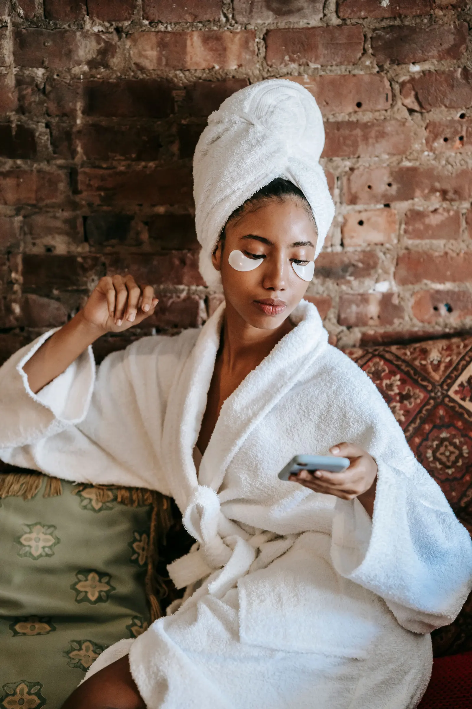 A woman in a towel wrap and white robe with under-eye patches, glancing at her phone