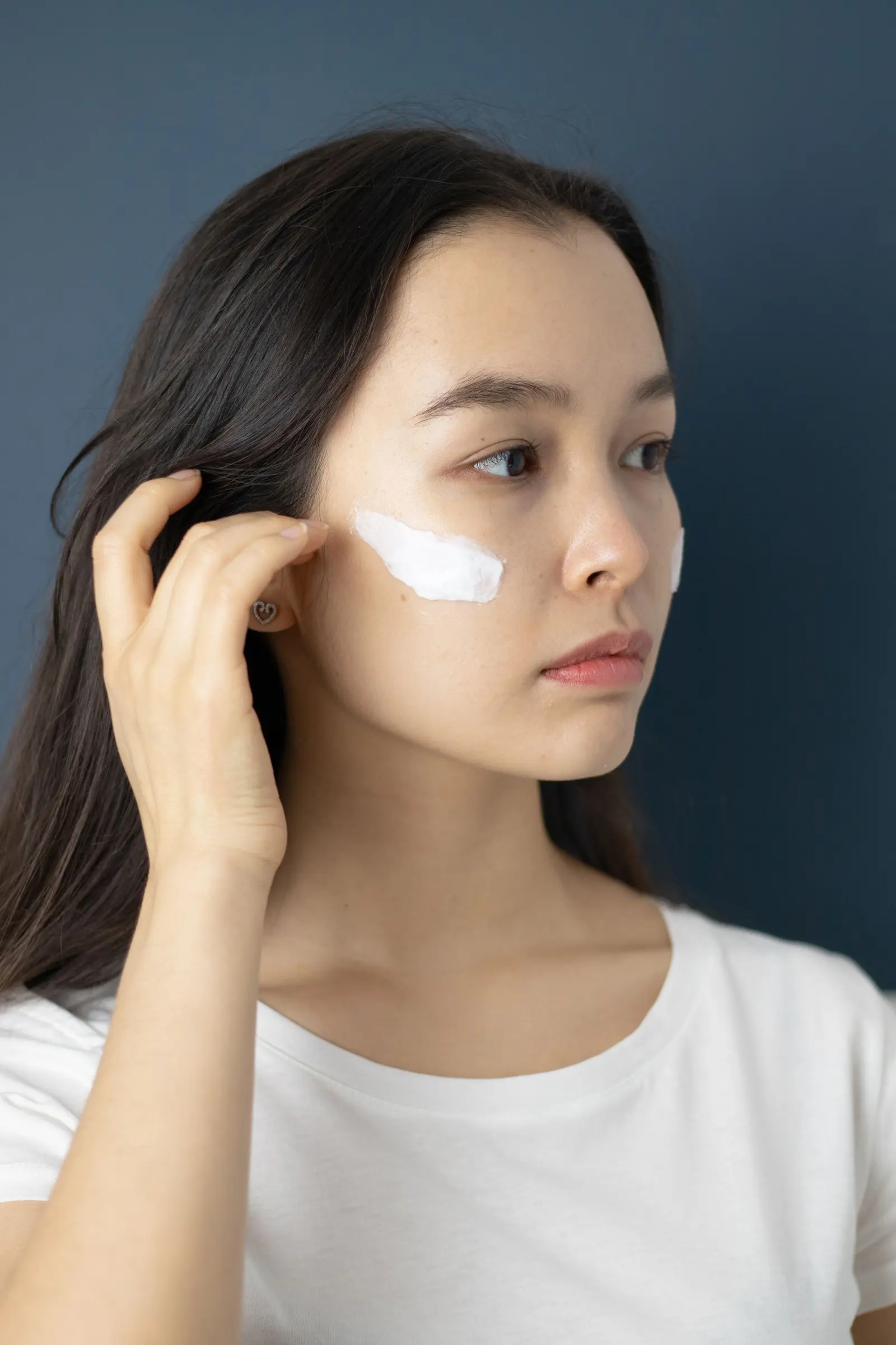 A woman with dark hair smoothing white cream onto her cheek against a navy wall