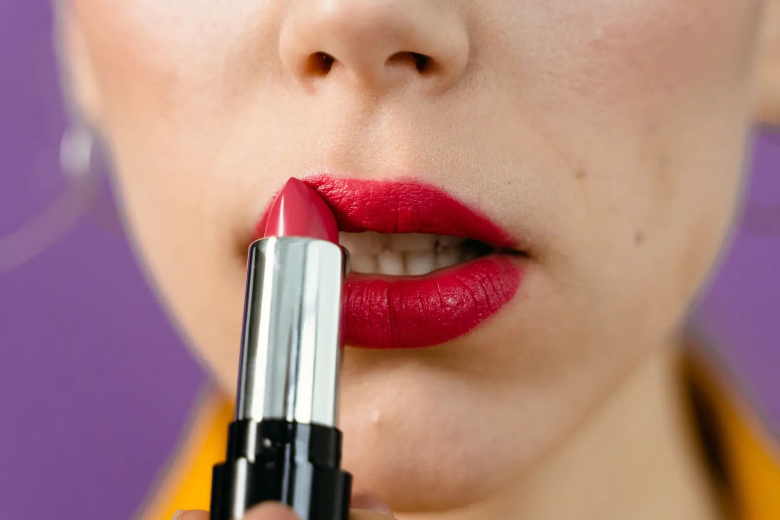 Close-up of a red lipstick being applied to a woman's lips against a purple backdrop